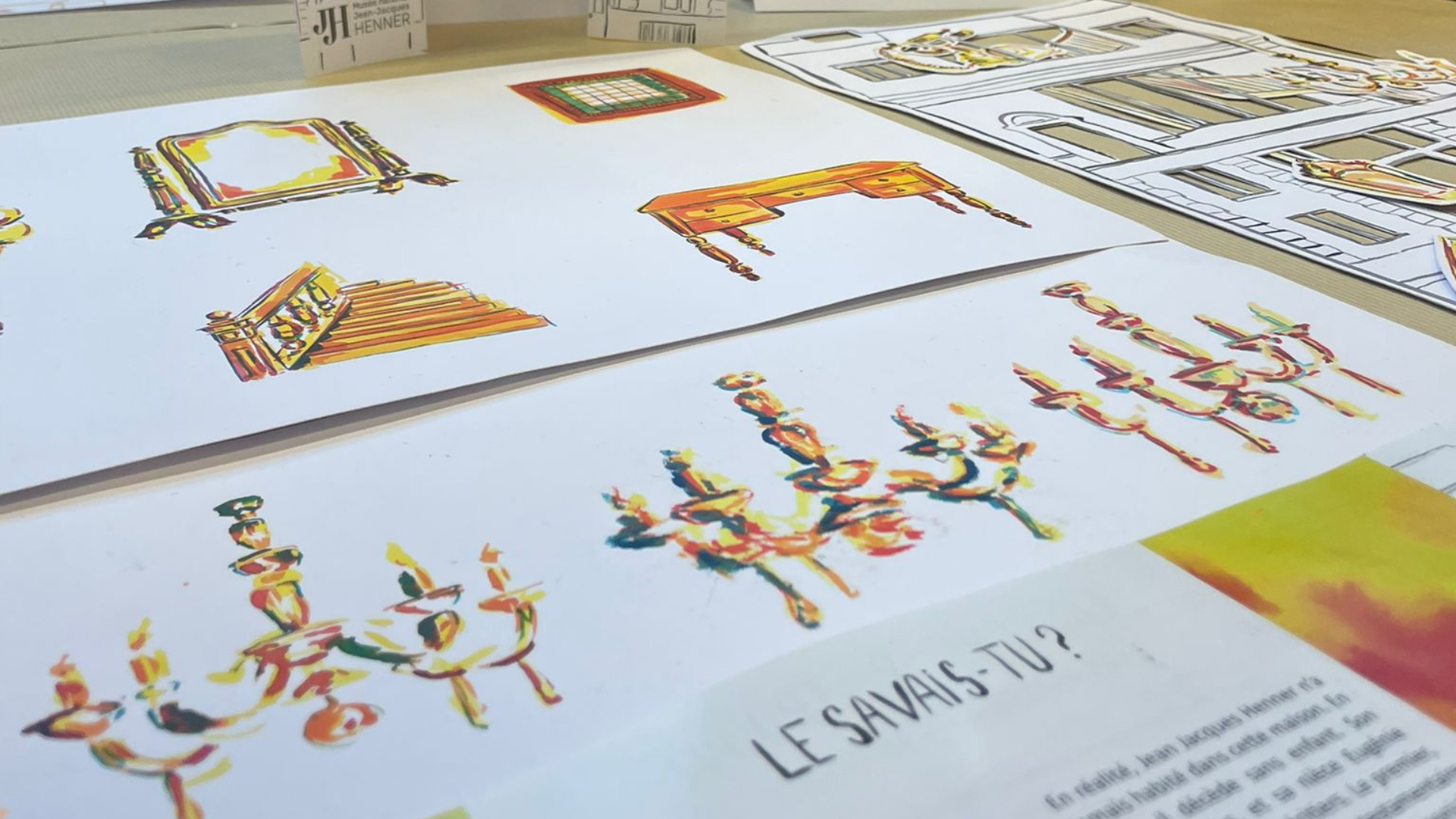 Planches d'illustrations colorées étalées sur une table, montrant des dessins de personnages et ornements avec le titre "LE SAVAIS-TU ?" visible.