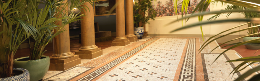 Salle de musée avec colonnes, mosaïques au sol, plantes vertes et lumière naturelle filtrant par le toit vitré.